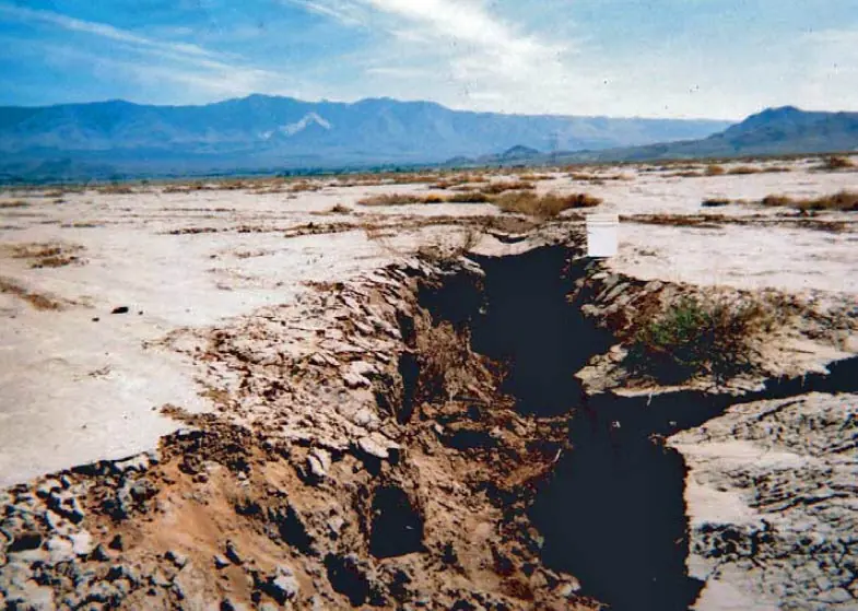 A wide ground fissure crossing dry terrain near Lucerne Lake, with a white bucket beside it for scale