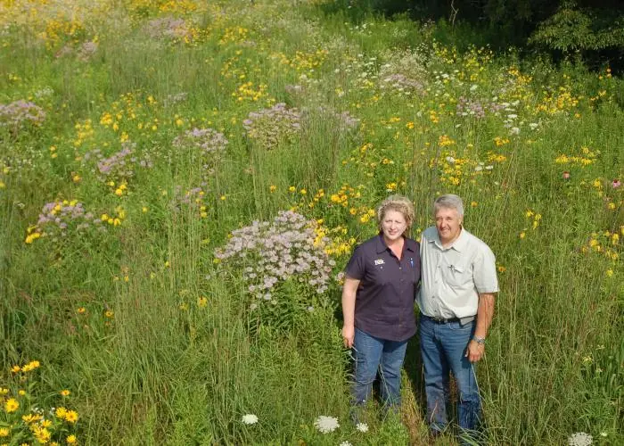Becky and Paul Rogers standing among tall grasses and purple, yellow and white wildflowers in Michigan.