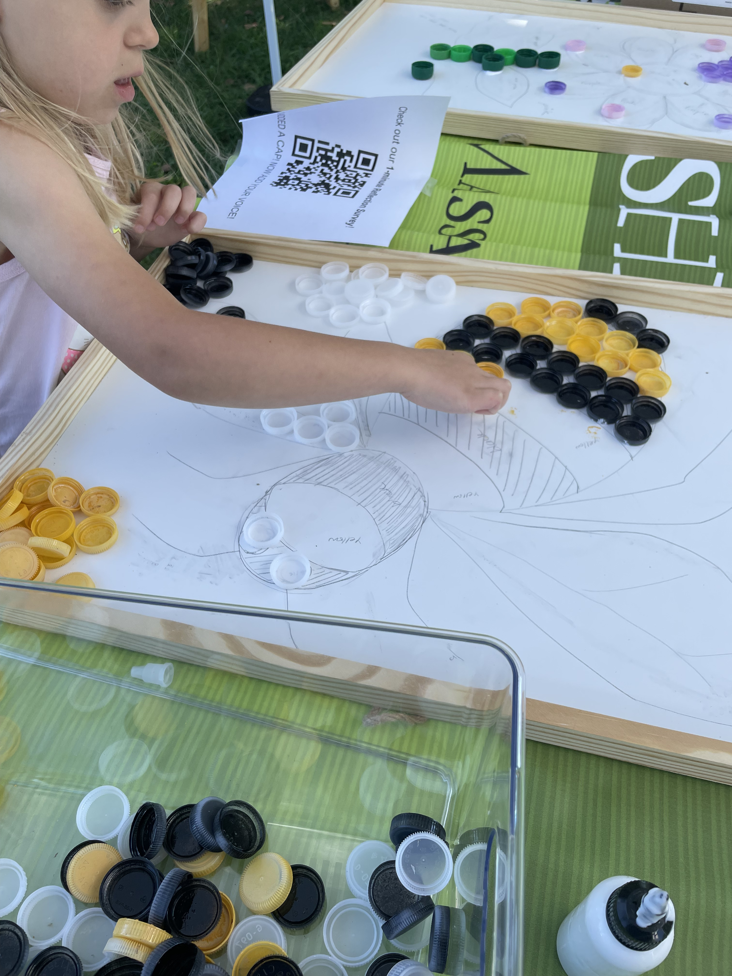 A child placing yellow and black bottle caps onto a drawn bee mosaic with bins of sorted caps nearby