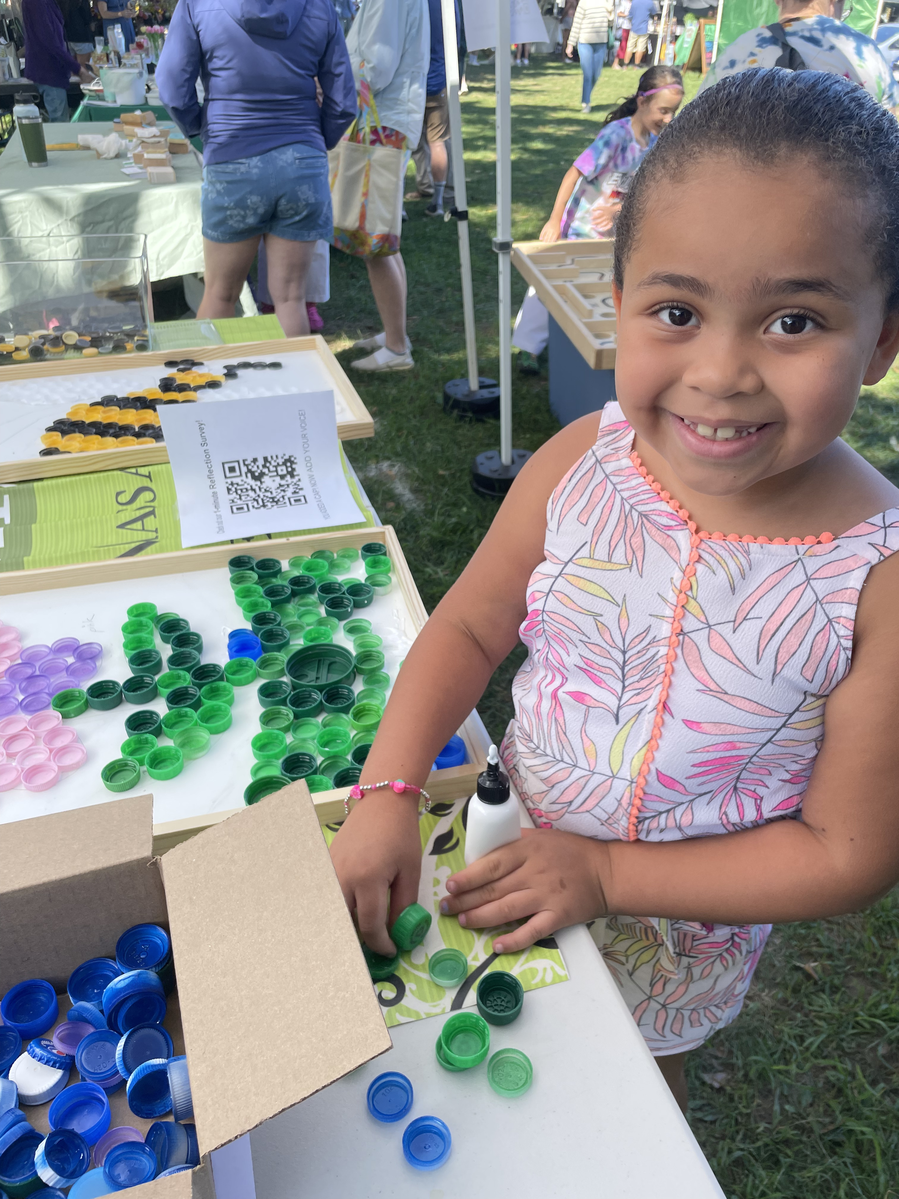 A smiling child placing green bottle caps onto a mosaic board at the sustainability booth