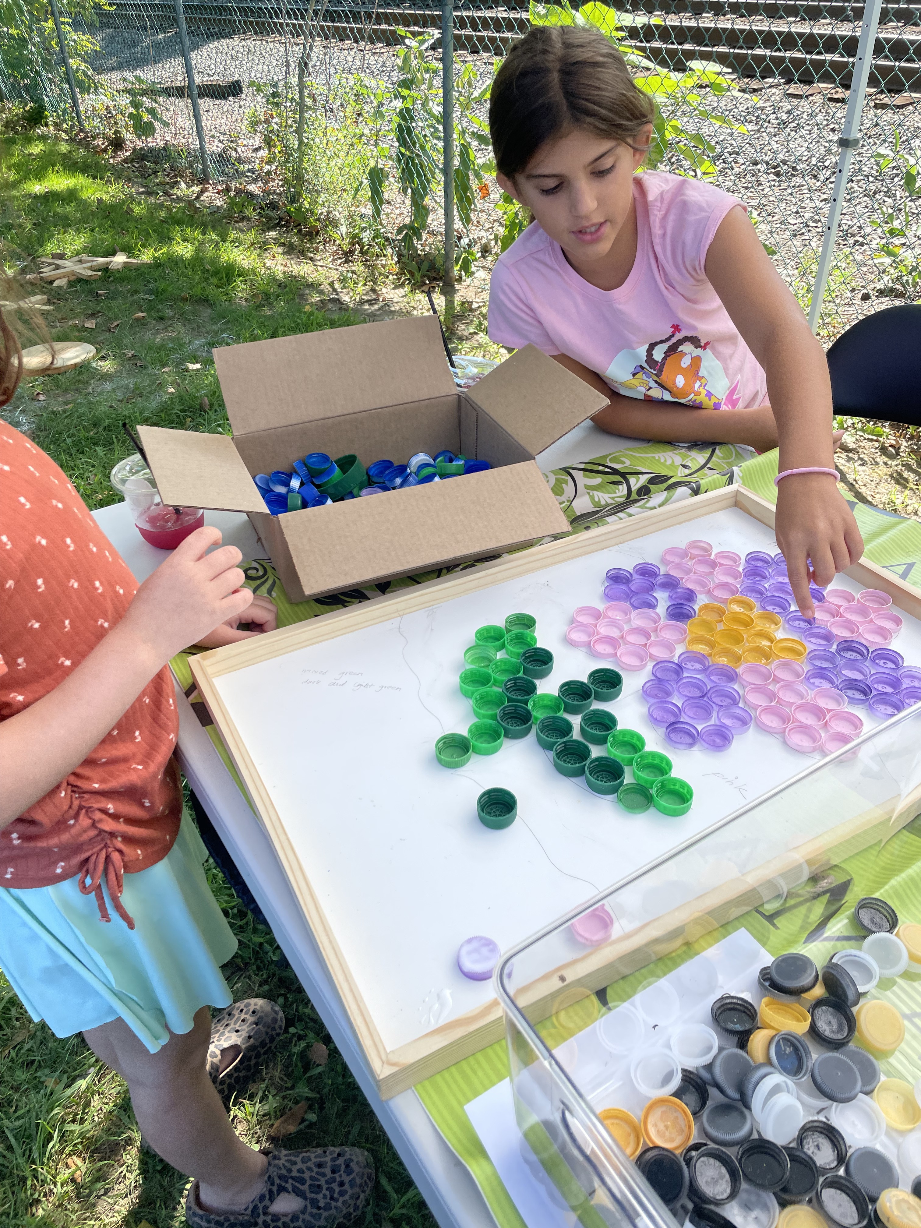Children sorting green, pink, and purple bottle caps into a flower mosaic with a box of blue caps